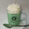 Ručna sveća u obliku matcha kafe sa kremastim vrhom, u staklenoj šolji sa logom Zdrava Kozmetika stavljena na tacnu sa kasikom pored.