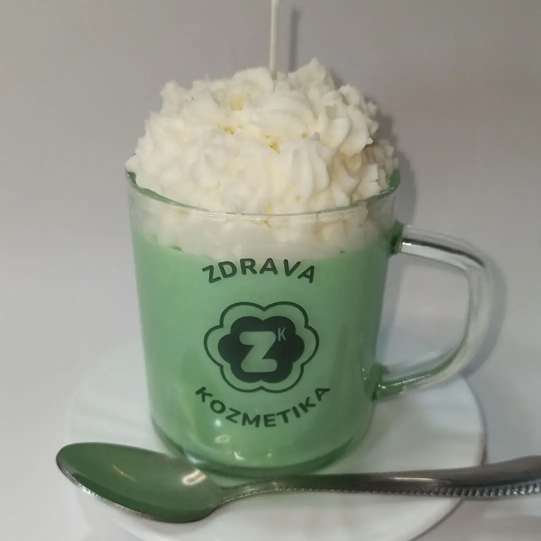 Ručna sveća u obliku matcha kafe sa kremastim vrhom, u staklenoj šolji sa logom Zdrava Kozmetika stavljena na tacnu sa kasikom pored.