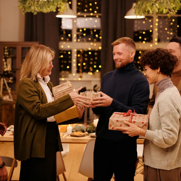 Grupa ljudi se okuplja u toplom, praznično ukrašenom domu, razmenjujući umotane poklone dok se smeju. Svetlucave lampice krase pozadinu.